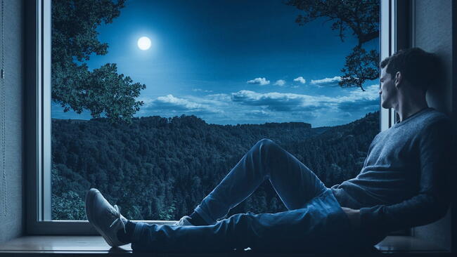 Man laying on a windowsill, looking out into the night sky and a darkened forest