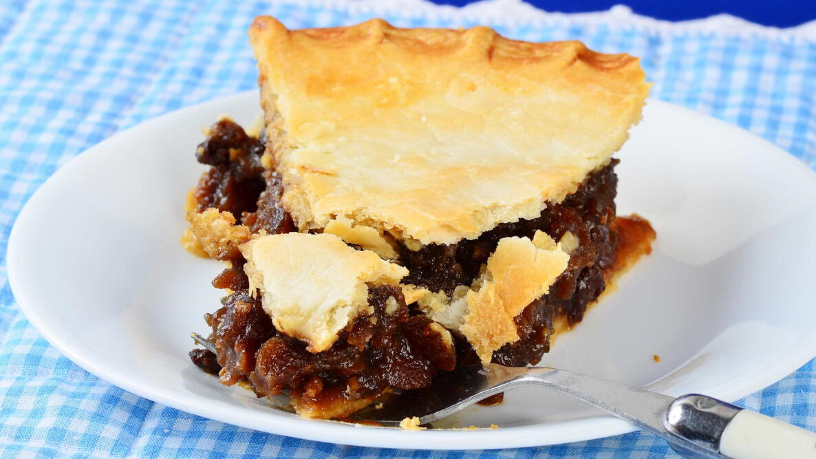 Bite cut from flaky crust of old-fashioned mincemeat pie on fork in country setting on blue gingham place mat.