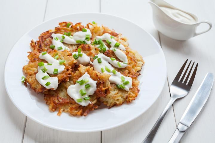 potato latkes with sour cream and chives on a white plate on a white wooden table with a fork and knife